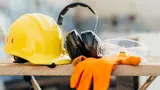 A yellow hard hat, black headphones, and orange gloves with clear safety glasses sitting on a flat surface.